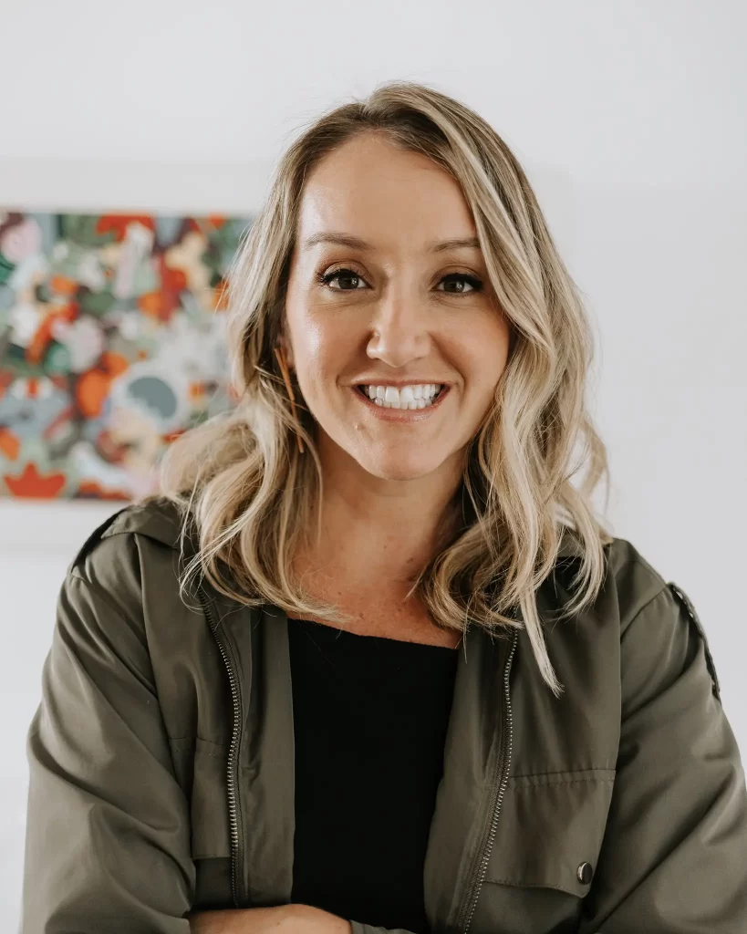 Photo of Jessica Johnson, co-founder of Rosyfinch: Jess is smiling, transmitting sympathy and confidence. She is wearing an olive-green jacket and is in an environment illuminated by a colorful work of art in the background, highlighting her relaxed and professional style.